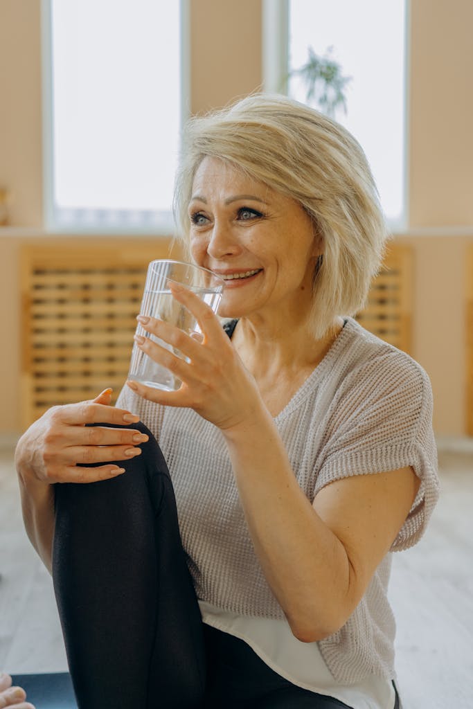 A smiling woman with blond hair holds a glass while sitting indoors, radiating warmth and positivity.