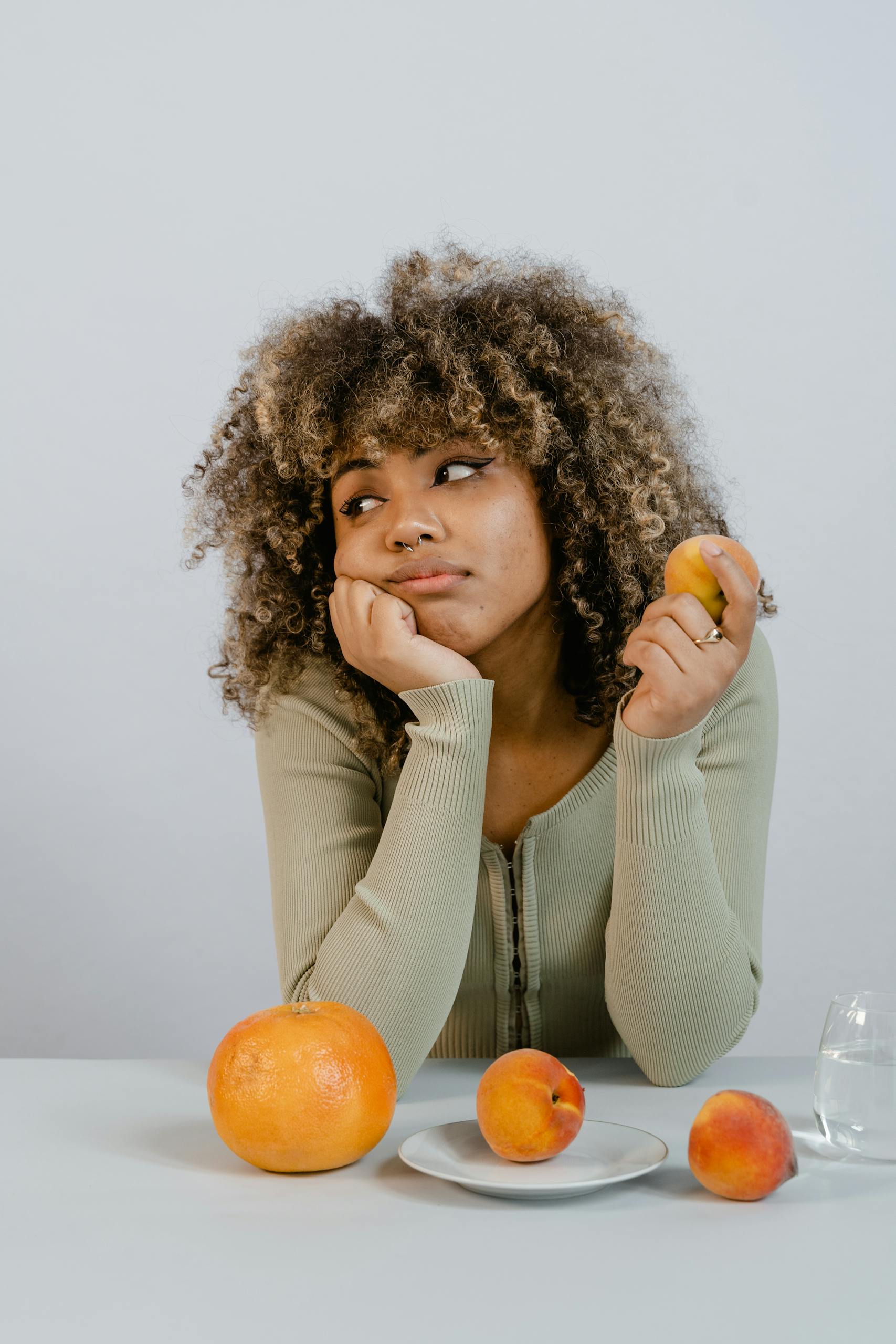 A thoughtful woman with curly hair contemplates holding a peach with other fruits on a table.