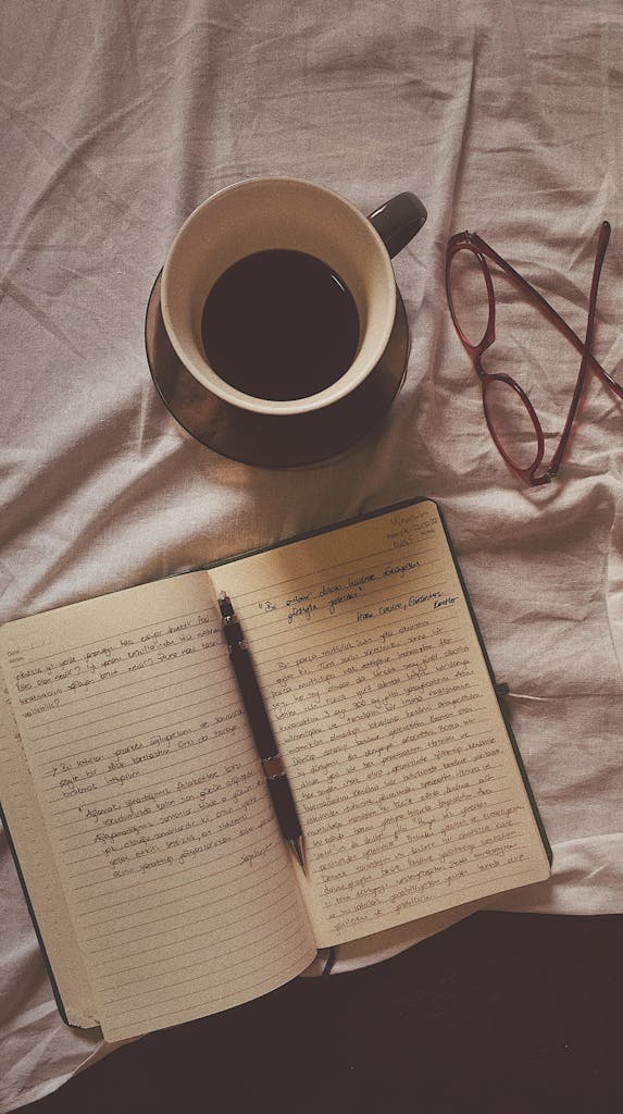 Flat lay of coffee, open notebook, pen, and eyeglasses on a fabric surface.
